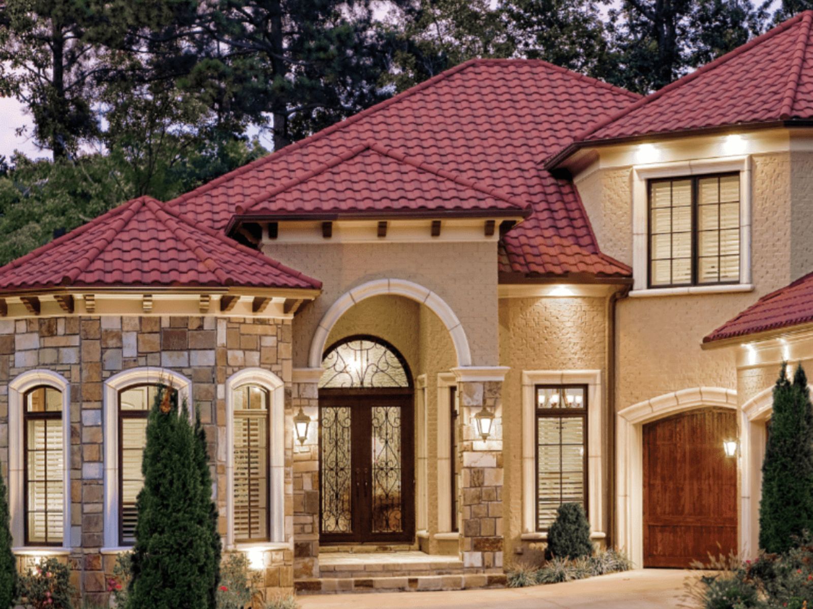 Luxury residential home with a red clay tile roof, stone and stucco exterior, arched entryway, and warm exterior lighting surrounded by trees.
