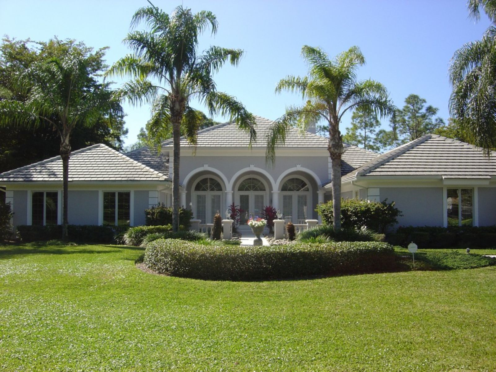Front-facing view of a Mediterranean-style home with a light gray concrete tile roof, symmetrical hip rooflines, arched windows, and palm trees framing a manicured lawn and landscaped entry.