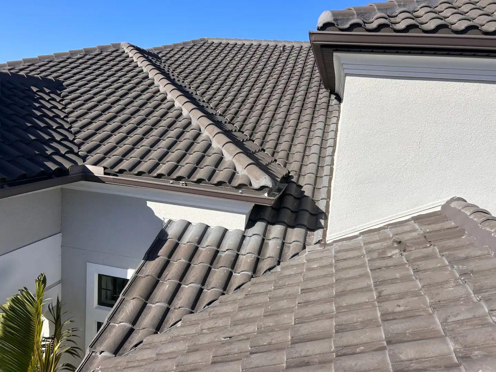 Close-up view of a residential home with a gray concrete tile roof, intersecting rooflines, and stucco exterior under a clear blue sky.