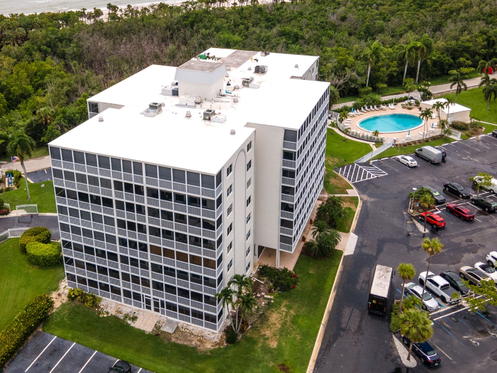 Aerial view of a mid-rise residential building with a white flat roof, surrounding parking lot, and nearby swimming pool bordered by trees