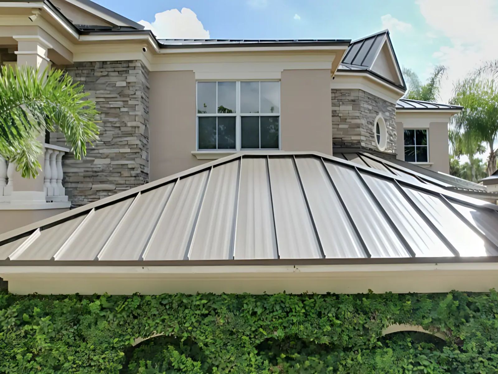 A front-facing view of a modern residential home featuring a light gray standing-seam metal roof over a covered entry, with crisp vertical seams, stone-accented exterior walls, white trim, and lush greenery below under a bright blue sky.