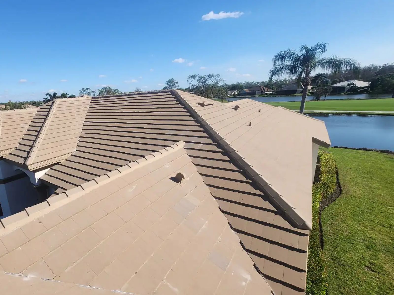 An elevated view of a tan concrete tile roof on a residential home, showing multiple roof planes with clean ridgelines and uniform tile spacing, set against a landscaped backyard with a pond, palm trees, and a clear blue sky.