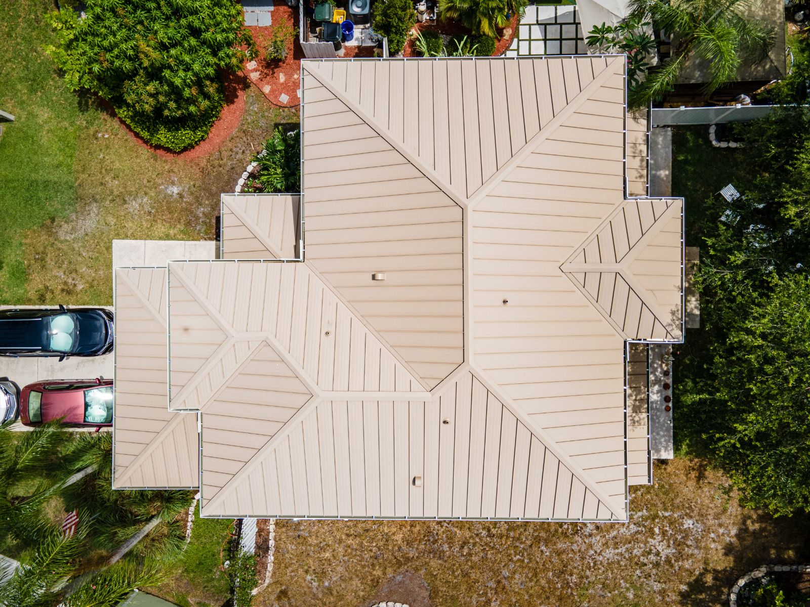 Aerial top-down view of a light beige standing-seam metal roof on a residential home, showing multiple roof planes, crisp seams, clean geometry, and surrounding landscaped yard with palm trees.